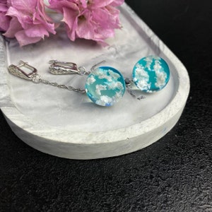 Puede incluir: Un par de pendientes colgantes de plata con abalorios redondos de resina azul con nubes blancas. Los pendientes est&aacute;n sobre una superficie de m&aacute;rmol blanco.