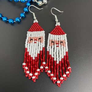 May include: Handmade beaded Santa Claus earrings. The earrings feature a red hat, white beard, and a pink face. The earrings have a fringe of red and white beads. The earrings are made with silver-colored ear hooks.