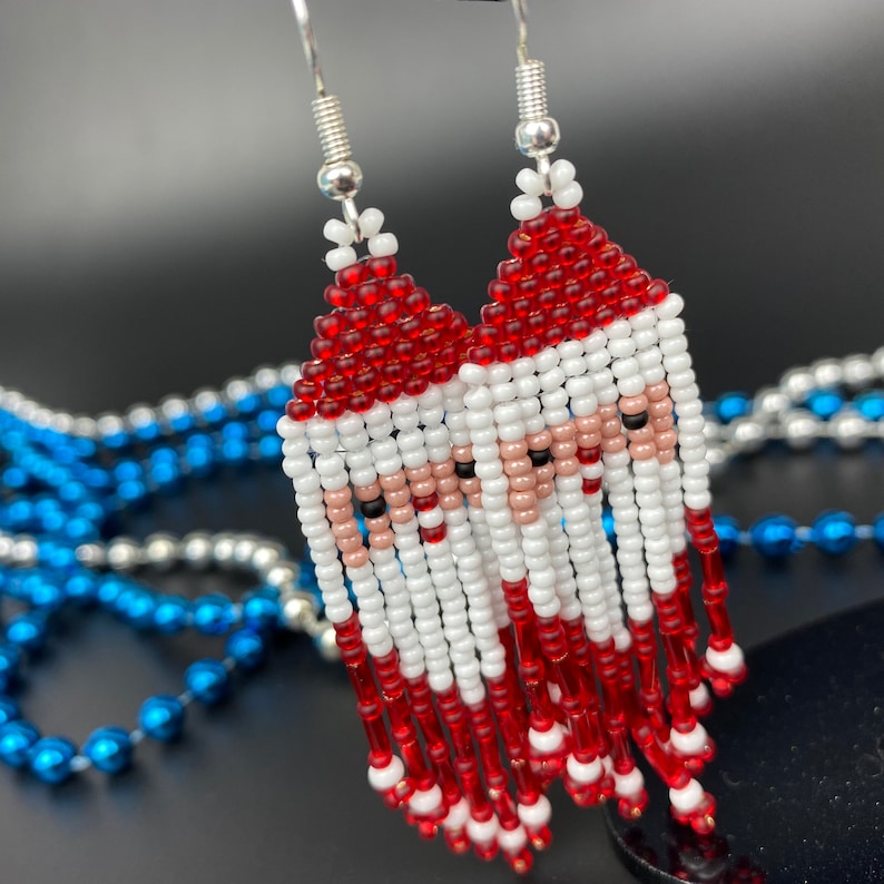 May include: Handmade beaded earrings featuring a Santa Claus design. The earrings have a red hat, white beard, and a skin-tone face. The earrings are made with small beads and have silver-colored ear wires. The background is dark.