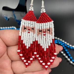 Peut inclure: Boucles d'oreilles faites à la main avec un motif de Père Noël. Les boucles d'oreilles sont composées de perles rouges, blanches, noires et beiges. Le bonnet du Père Noël est rouge et la barbe est blanche. Un accessoire festif pour les fêtes.