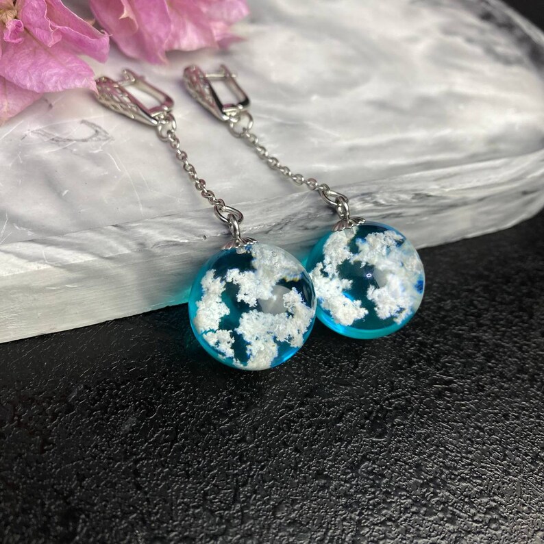 Puede incluir: Un par de pendientes de plata con colgantes de esferas de resina azul y blanca. Las esferas tienen un dise&ntilde;o nublado con motas blancas en toda la resina azul.