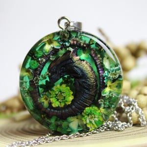Peut inclure: Un collier pendentif rond avec une chaîne argentée. Le pendentif est en résine transparente avec un motif sombre en spirale et des accents floraux verts et jaunes. Le pendentif mesure environ 3,8 cm de diamètre.