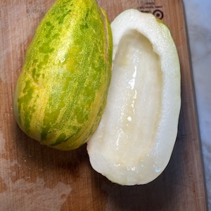 May include: A green and yellow cucumber cut in half, revealing the white flesh inside. The cucumber is on a wooden cutting board.