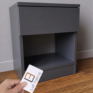 Pode incluir: Uma mesa de cabeceira cinza escuro com uma gaveta e uma prateleira aberta. A mesa de cabeceira é feita de madeira e tem um design moderno. Um cartão de visita com o logotipo "Loft Fabriq" é segurado em frente à mesa.
