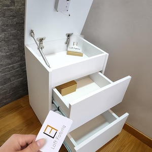 Puede incluir: Un mueble blanco con dos cajones abiertos y una tapa que se levanta. El mueble tiene una pequeña caja dentro de uno de los cajones. Una tarjeta con el texto "Loft Fabrig" se sostiene frente al mueble.