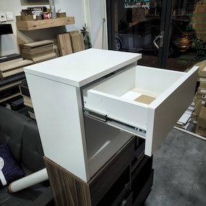 May include: A white bedside table with a single open drawer featuring metal side glides. The unit has a rectangular top surface and a clean, smooth finish. It is positioned atop a dark wood-toned cabinet with multiple storage compartments.