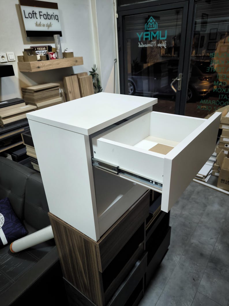 May include: A white bedside table with a single open drawer featuring metal side glides. The unit has a rectangular top surface and a clean, smooth finish. It is positioned atop a dark wood-toned cabinet with multiple storage compartments.