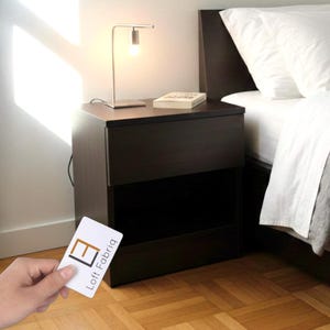 Pode incluir: Uma mesa de cabeceira castanha escura com duas gavetas, uma pequena lâmpada prateada e um livro em cima. Uma mão segura um cartão com o texto "Loft Fabriq". A mesa de cabeceira está ao lado de uma cama com roupa de cama branca.