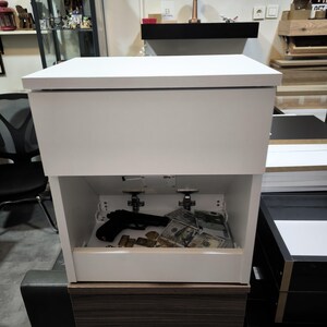 May include: A white bedside table with a rectangular top and a hidden storage compartment. The open lower section reveals a black handgun, several gold-colored bars, and a stack of one hundred dollar bills resting on the interior base surface.