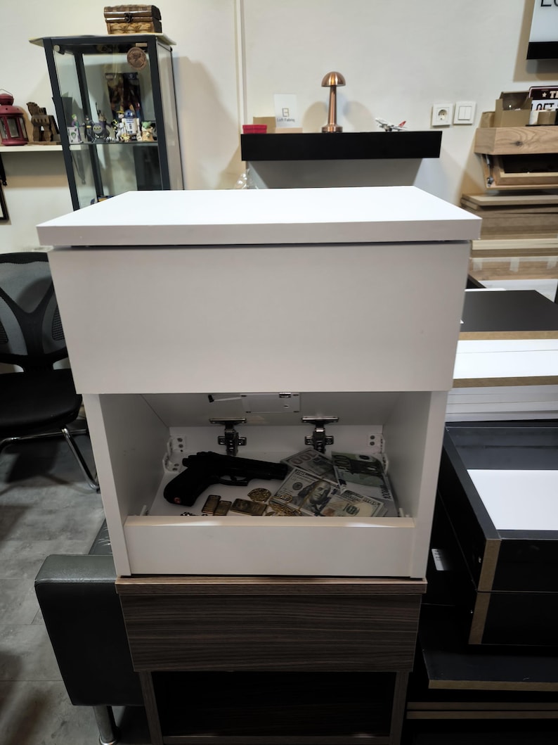 May include: A white bedside table with a rectangular top and a hidden storage compartment. The open lower section reveals a black handgun, several gold-colored bars, and a stack of one hundred dollar bills resting on the interior base surface.
