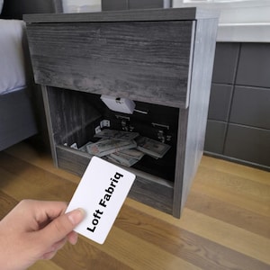 Pode incluir: Uma mesa de cabeceira cinza de madeira com um compartimento oculto cheio de dinheiro. A mesa de cabeceira tem uma gaveta e um compartimento inferior. Um cartão branco com o texto "Loft Fabriq" é segurado em frente à mesa de cabeceira.
