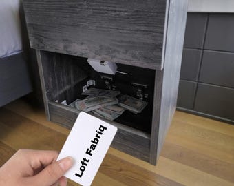 Può includere: Un comodino in legno grigio scuro con uno scomparto nascosto dotato di un meccanismo di chiusura elettronico bianco. All'interno dello spazio aperto sono visibili mazzette di banconote. Una carta bianca con il testo Loft Fabriq.