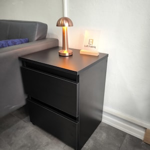 Pode incluir: Uma mesa de cabeceira preta com duas gavetas, encimada por uma lâmpada em forma de cogumelo cor de cobre e um letreiro que diz "Loft Fabriq". A lâmpada está acesa, emitindo um brilho quente. A mesa de cabeceira está ao lado de um sofá cinza.