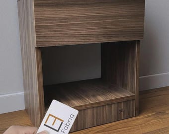 Può includere: Un comodino in legno marrone con un cassetto e un ripiano aperto. Il comodino ha un motivo a venature del legno e un piccolo asterisco decorativo sul lato. Un biglietto da visita con "Loft Fabriq" è tenuto davanti al comodino.