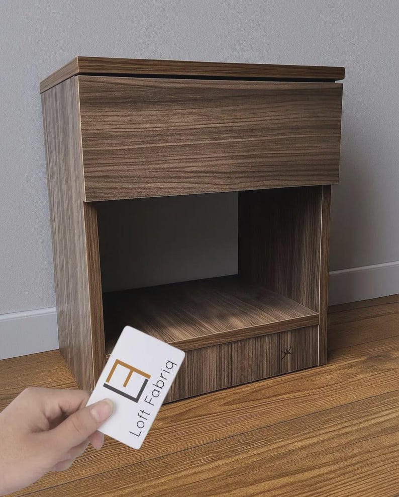 May include: A brown wooden nightstand with a drawer and an open shelf. The nightstand has a wood grain pattern and a small, decorative asterisk on the side. A business card with the text "Loft Fabriq" is held in front of the nightstand.