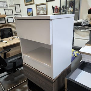 May include: A white bedside table with a single top drawer and an open storage shelf below. The unit features a smooth, matte finish and a rectangular structure, positioned on top of a dark wood-toned cabinet base in a workshop environment.