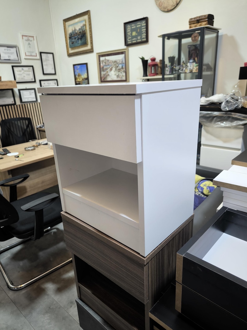 May include: A white bedside table with a single top drawer and an open storage shelf below. The unit features a smooth, matte finish and a rectangular structure, positioned on top of a dark wood-toned cabinet base in a workshop environment.