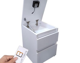 May include: A white bedside table with two drawers and a top compartment that opens upward using a gas strut mechanism. A black electronic lock is mounted on the interior of the lid. A white card with the text Loft Fabriq is visible.