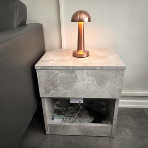Pode incluir: Uma pequena mesa de cabeceira cinza com efeito mármore com uma lâmpada cor de cobre em cima. A lâmpada está acesa, emitindo um brilho quente. A mesa de cabeceira tem uma prateleira aberta que revela dinheiro e uma arma.