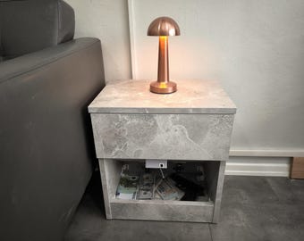 Può includere: Un piccolo comodino grigio effetto marmo con una lampada color rame sopra. La lampada è accesa, emettendo un bagliore caldo. Il comodino ha un ripiano aperto che rivela contanti e una pistola.