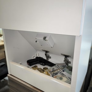 May include: A white cabinet with a hidden compartment featuring a downward-opening door. Inside the compartment, there is a black handgun, a stack of one hundred dollar bills, and gold-colored items resting on the flat interior surface.