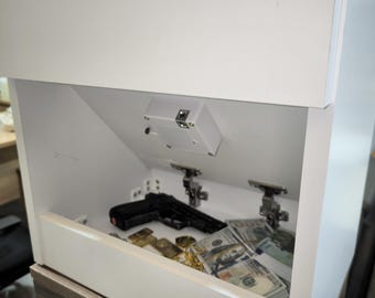 May include: A white cabinet with a hidden compartment featuring a downward-opening door. Inside the compartment, there is a black handgun, a stack of one hundred dollar bills, and gold-colored items resting on the flat interior surface.