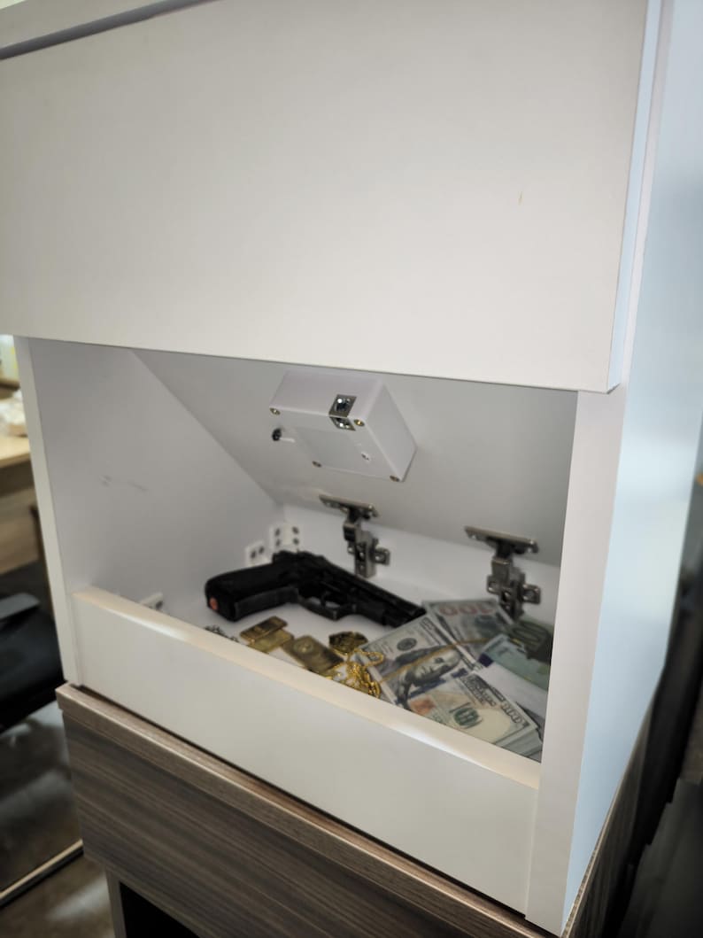 May include: A white cabinet with a hidden compartment featuring a downward-opening door. Inside the compartment, there is a black handgun, a stack of one hundred dollar bills, and gold-colored items resting on the flat interior surface.