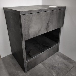 Pode incluir: Um armário cinza moderno com um acabamento texturizado. O armário tem uma gaveta e uma prateleira aberta abaixo. O design é minimalista e o armário está colocado em um canto.