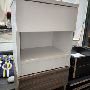 May include: A white bedside table with a flat top surface and an open storage compartment below. The unit features a clean, rectangular design with smooth panels and a solid construction, providing a simple and functional storage solution.