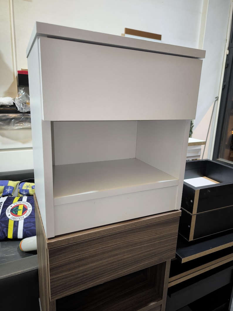 May include: A white bedside table with a flat top surface and an open storage compartment below. The unit features a clean, rectangular design with smooth panels and a solid construction, providing a simple and functional storage solution.