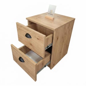 May include: A light brown wood grain bedside cabinet featuring two open drawers with black semi-circular handles and metal sliding tracks. A small rectangular sign with the text Loft Fabriq is positioned on the top surface of the furniture piece.