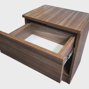 May include: A bedside table with a dark brown wood grain finish featuring a single pull-out drawer. The drawer front matches the wood grain pattern of the frame, while the interior of the drawer is finished in a solid white color.