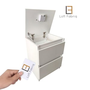 Op de afbeelding: Een wit nachtkastje met twee lades en een bovenste compartiment dat naar boven opent met metalen scharnieren. De unit is voorzien van een klein elektronisch slotmechanisme aan de onderkant van het deksel. Het Loft Fabriq logo is zichtbaar op de kaart.