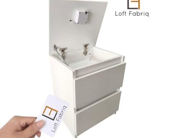 Può includere: Un comodino bianco con due cassetti e uno scomparto superiore che si apre verso l'alto su cerniere metalliche. L'unità è dotata di un piccolo meccanismo di chiusura elettronica sul lato inferiore del coperchio. Il logo Loft Fabriq è visibile.