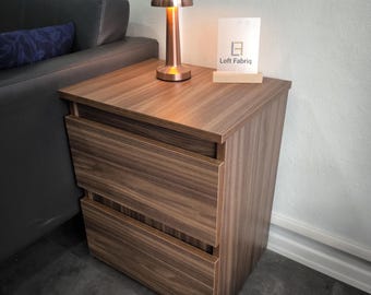 May include: A wooden bedside cabinet with two drawers featuring a horizontal wood grain pattern. On top sits a metallic copper-coloured mushroom-shaped lamp and a white rectangular sign with the text Loft Fabriq printed in black.