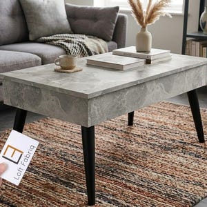 Könnte beinhalten: Ein rechteckiger Couchtisch mit grauer Kunstmarmor-Optik und vier konisch zulaufenden schwarzen Beinen. Der Tisch verfügt über eine Hubtischplatte und wird mit einer weißen Visitenkarte mit der Aufschrift Loft Fabriq in Schwarz und Orange gezeigt.
