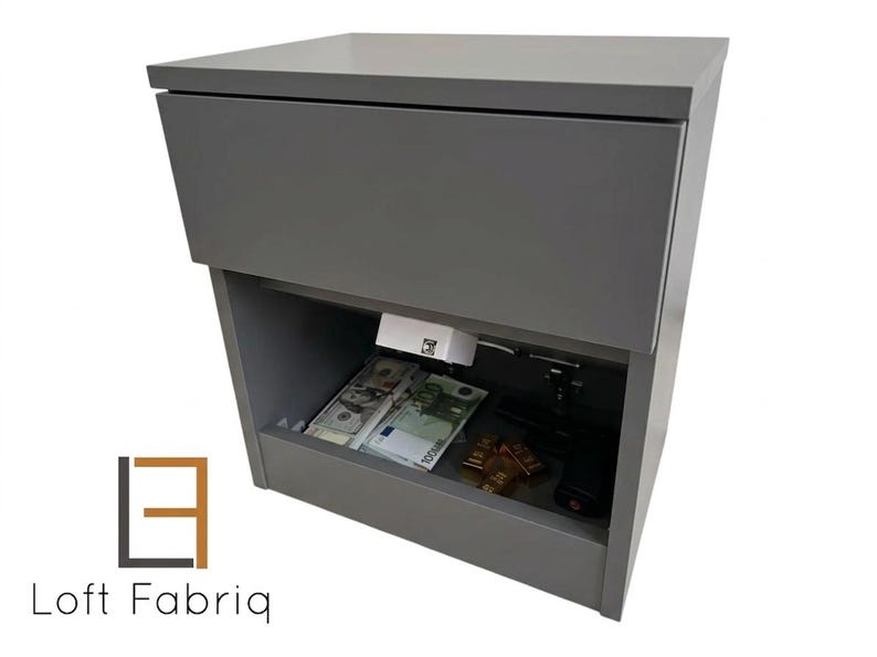 May include: A gray cabinet with a hidden compartment, containing cash, gold bars, and a handgun. The cabinet has a top drawer and an open lower section. The logo "Loft Fabriq" is visible on the bottom left.