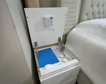 Può includere: Comodino bianco con due cassetti e un vano nascosto. Lo scomparto superiore è aperto, rivelando una cartella blu, denaro e altri oggetti. Il comodino è accanto a un letto con testiera color crema.