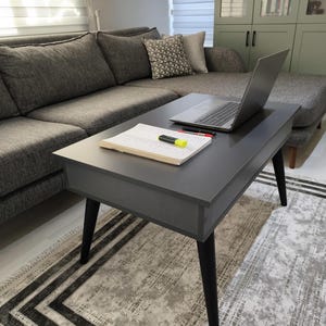 May include: A dark gray rectangular coffee table with four tapered black legs. On the tabletop sits an open spiral-bound notebook with a yellow highlighter, two pens, and a silver laptop computer positioned toward the back of the surface.