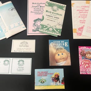 May include: A collection of Cabbage Patch Kids items including a birth certificate, adoption papers, a guide to hairstyling, and a doll with a pink and white outfit. The doll is wearing a headband with a bow and has a curly hairstyle.