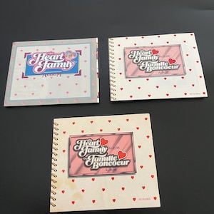 May include: Three spiral bound photo albums with a pink and white heart pattern. The covers feature the text "The Heart Family" and "La Famille Boncoeur" with a red heart graphic.