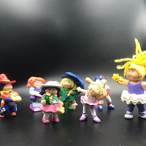 May include: A collection of plastic figurines of the Cabbage Patch Kids characters. The characters are dressed in various outfits, including a cowboy hat, a ballerina dress, and a green dress with a matching umbrella.