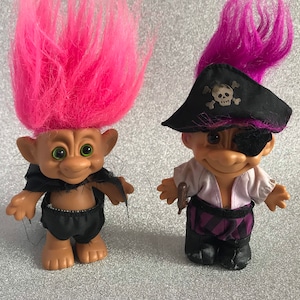May include: Two troll dolls dressed as pirates. One troll has pink hair and a black cape, the other has purple hair and an eye patch. Both trolls are wearing black trousers and boots.