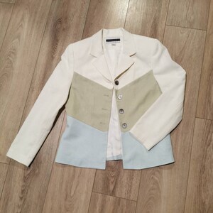 Puede incluir: Una chaqueta blanca con un diseño de bloques de color verde claro y azul claro. La chaqueta tiene cierre de botón y mangas largas.