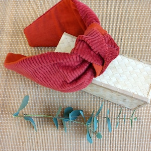 Op de afbeelding: Een rode corduroy hoofdband met een geknoopt ontwerp. De hoofdband heeft een levendige rode tint en lijkt gemaakt te zijn van een zachte, getextureerde stof. Het ligt op een geweven bamboemat, met een kleine, rechthoekige geweven doos.