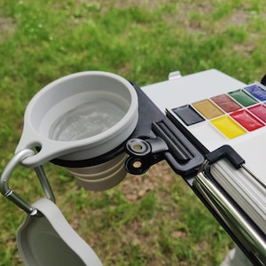 Może przedstawiać: Paleta do akwareli w kolorze czarnym i szarym z wbudowanym uchwytem na kubek z wodą. Paleta ma klips do mocowania do powierzchni i metalowy uchwyt. Kubek na wodę jest wykonany z silikonu i jest składany.