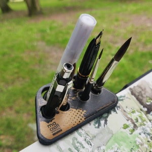 Peut inclure: Un porte-stylos noir et brun avec cinq emplacements pour stylos et pinceaux. Le porte-stylos porte le logo "ma craft workshop" et un motif de pois noir et blanc. Il contient un stylo noir, un stylo argenté, un stylo blanc et un pinceau brun.