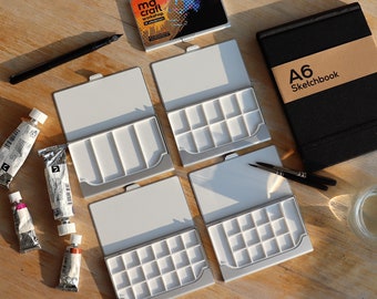 May include: Four silver watercolor palettes with white wells, each with a white lid.  The palettes are arranged in a row on a wooden surface.  There are five tubes of watercolor paint, a black pen, two paintbrushes, and a black sketchbook with the text "A6 Sketchbook" on the cover.