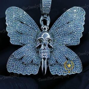 May include: A silver-toned pendant featuring a butterfly design with a skull at the center. The wings are encrusted with small, sparkling stones. The pendant has a bail for a chain. The overall design is intricate and detailed.