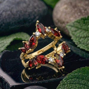 May include: A gold ring guard featuring marquise-cut deep red garnet gemstones and small round white accent stones arranged in a leaf-like pattern. The band is designed to frame another ring, showcasing a vibrant contrast between the red and gold.